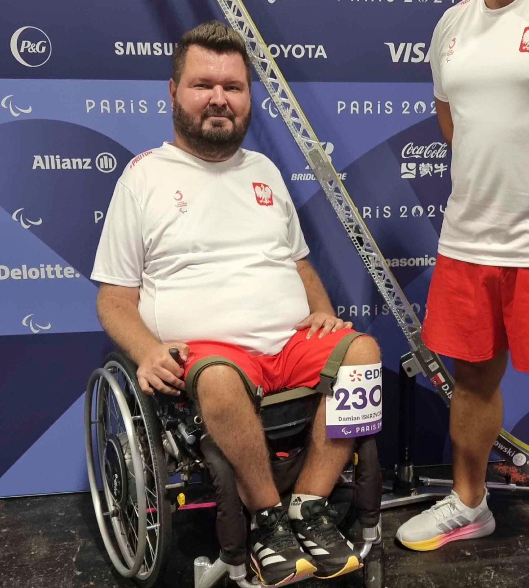 39-letni Damian Iskrzycki siedzi na wózku, ubrany w biało-czerwony sportowy strój reprezentacji Polski, z numerem startowym na kolanie. Obok widać fragment rampy do gry w boccię oraz uciętą sylwetkę mężczyzny również w biało-czerwonym stroju. Za nimi ścianka z logotypami Igrzysk Olimpijskich w Paryżu.