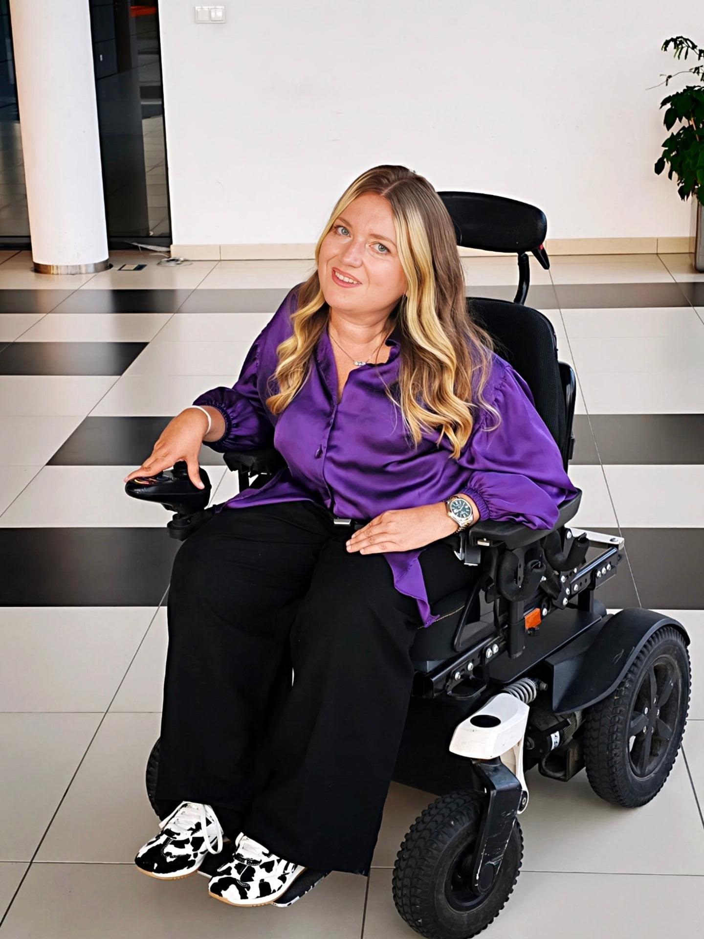Sylwia Błach - młoda kobieta na elektrycznym wózku, w błyszczącej fioletowej koszuli, czarnych spodniach i sportowych butach w zwierzęcy wzór. Włosy długie falowane