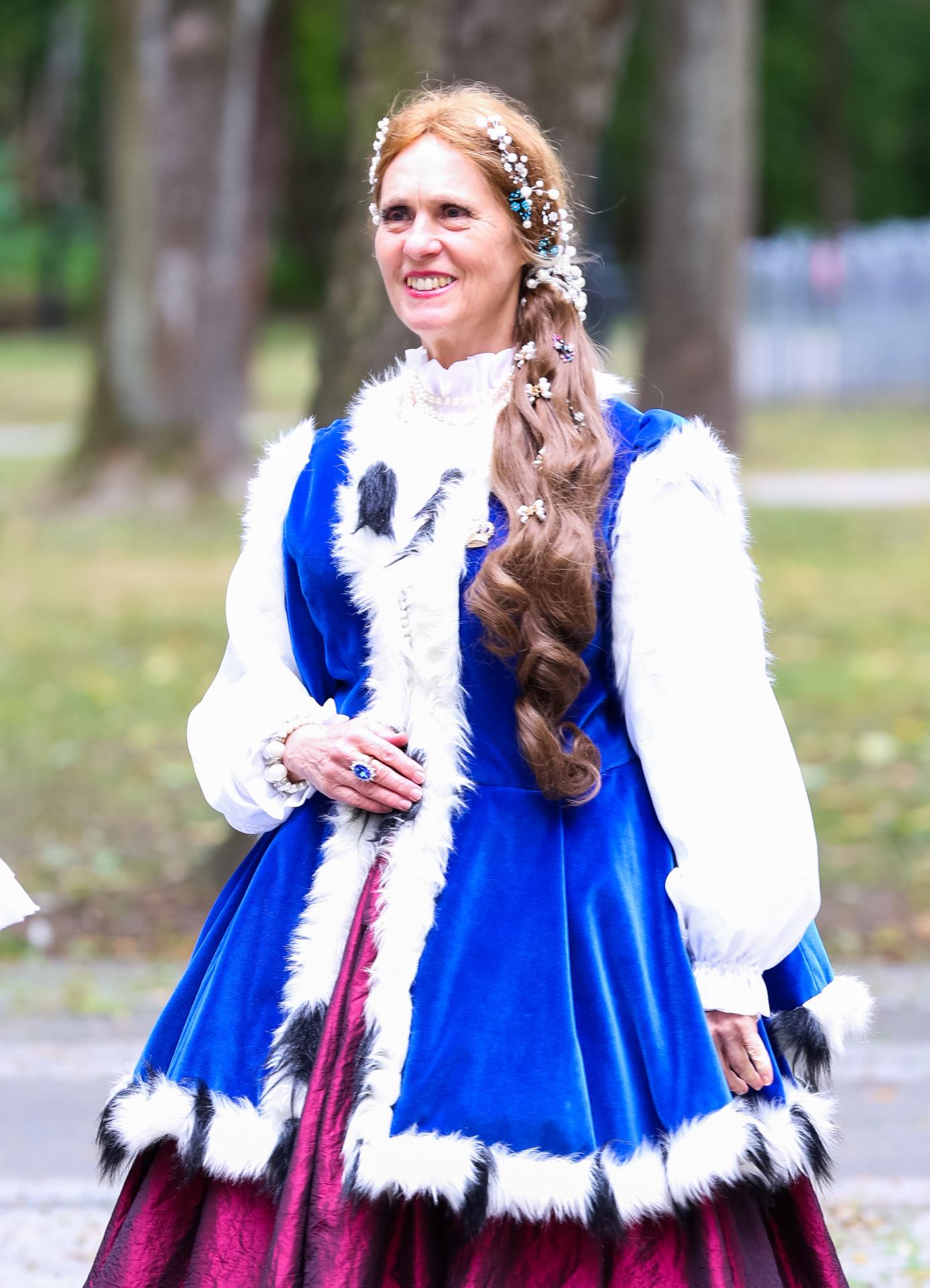 Maria Czerwińska w stroju kobiecym z XVIII wieku, w kolorze białym, indygo i purpurowym. Uśmiechnięta kobieta pozuje w parku.